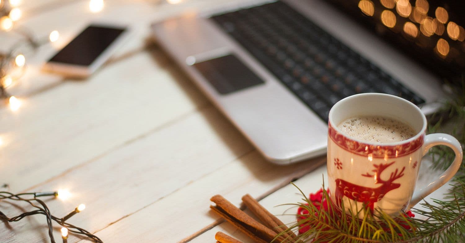 Holiday coffee mug next to laptop and phone surrounded by lights